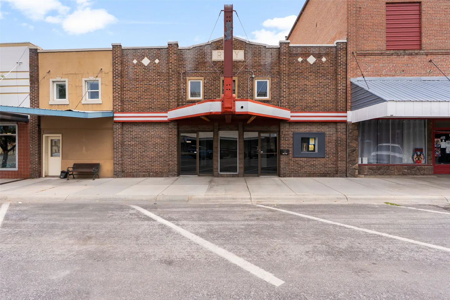 Exterior view of a brick building with a red and white awning over glass doors. "RY" is displayed above the awning. Parking spaces are in front.