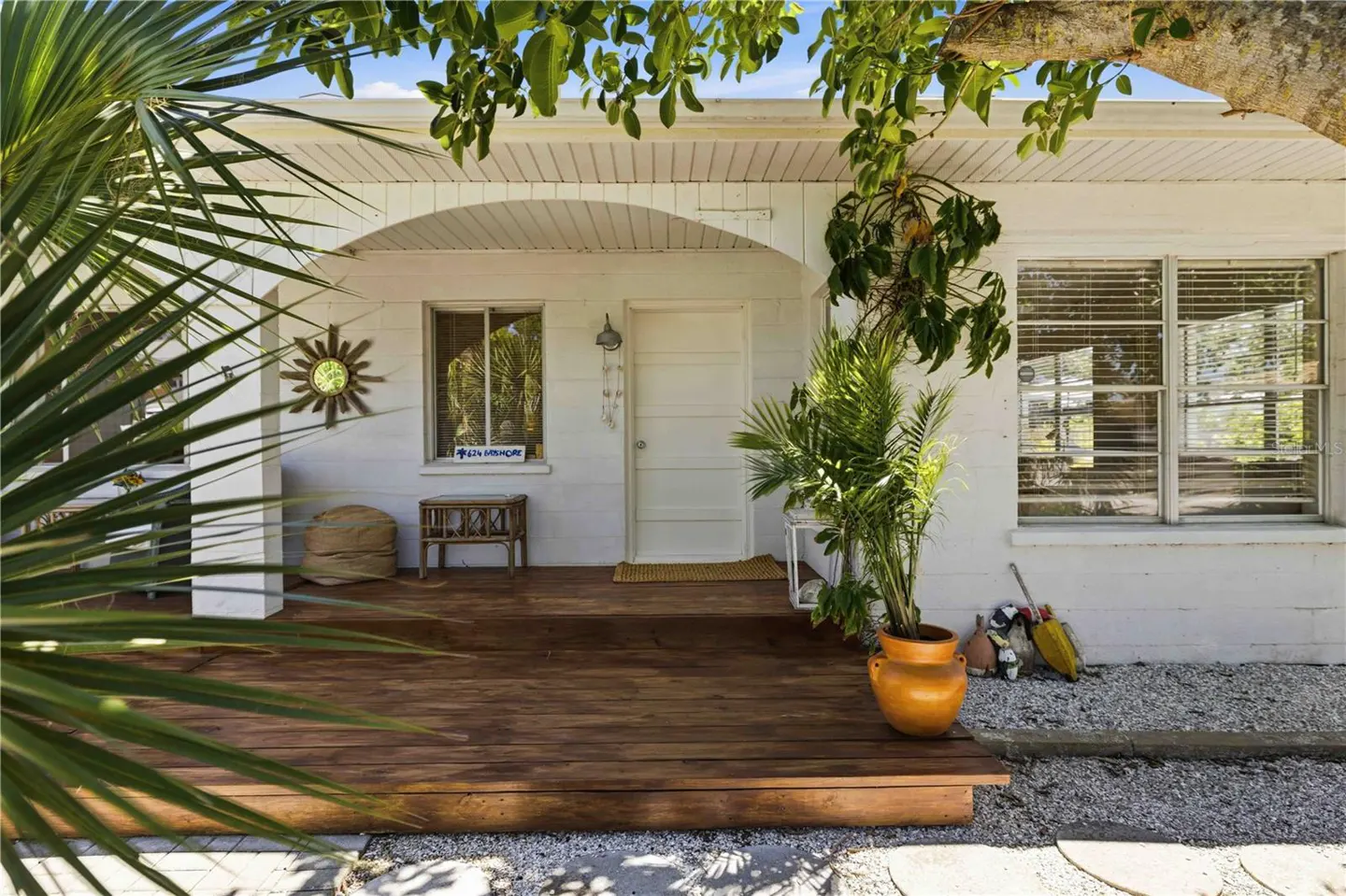 A white house with a wooden porch, a white door, and a window with blinds. A potted plant sits on the porch.