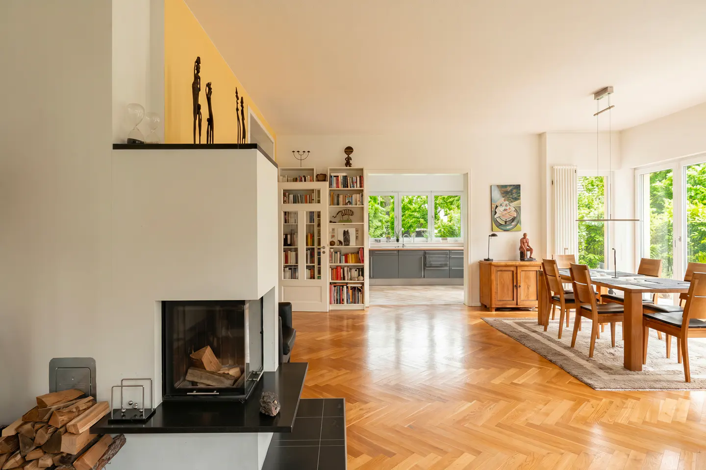 Open-plan living area with a fireplace, herringbone wood floors, dining table, and a view into a modern kitchen.