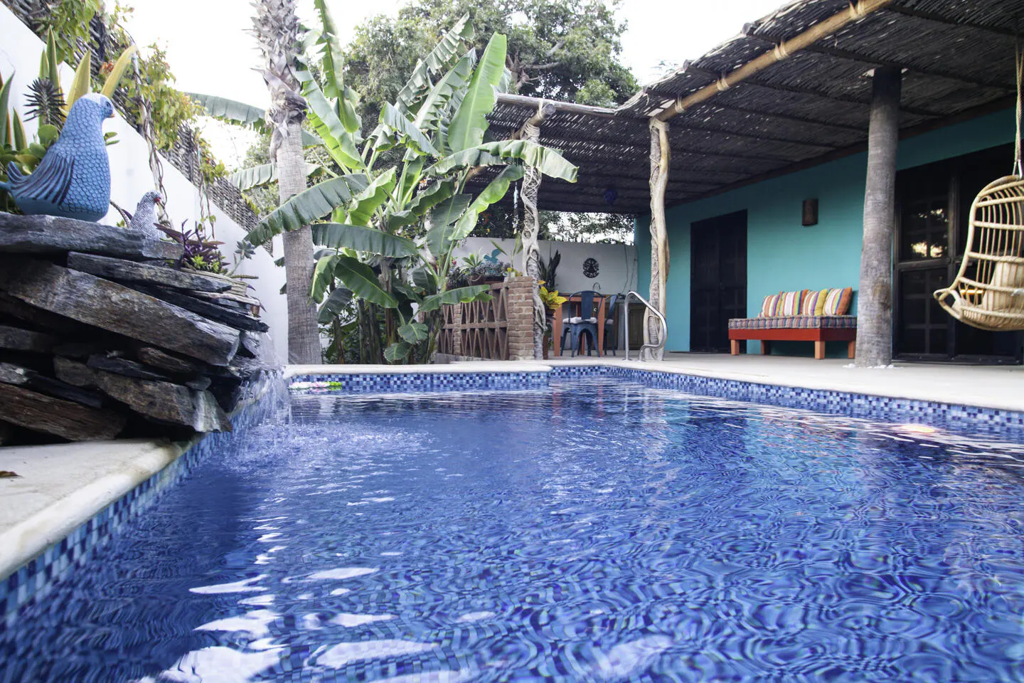 A bright blue pool with a waterfall feature, surrounded by lush greenery and a turquoise building with a thatched roof.