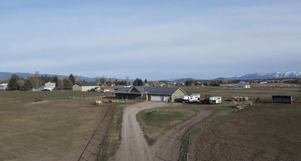 Aerial view of a ranch-style house with a gray roof, surrounded by fields, horses, and mountains in the background.