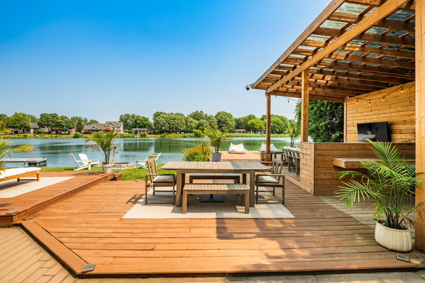 Outdoor wooden deck with dining table, chairs, and a covered lounge area overlooking a lake on a sunny day.