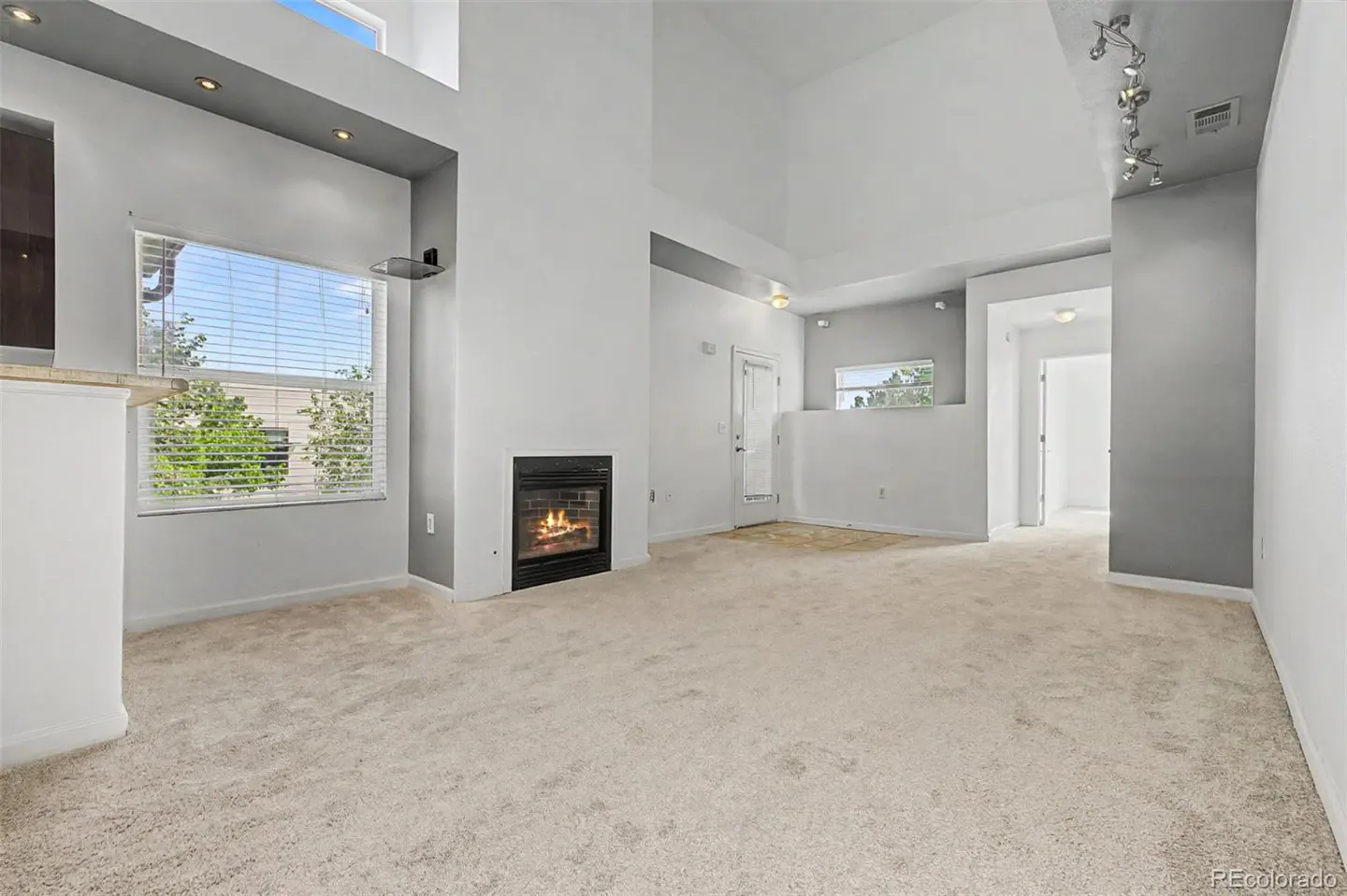 Bright, open living room with beige carpet, gray walls, and a black fireplace with a fire burning. A large window with blinds overlooks trees.