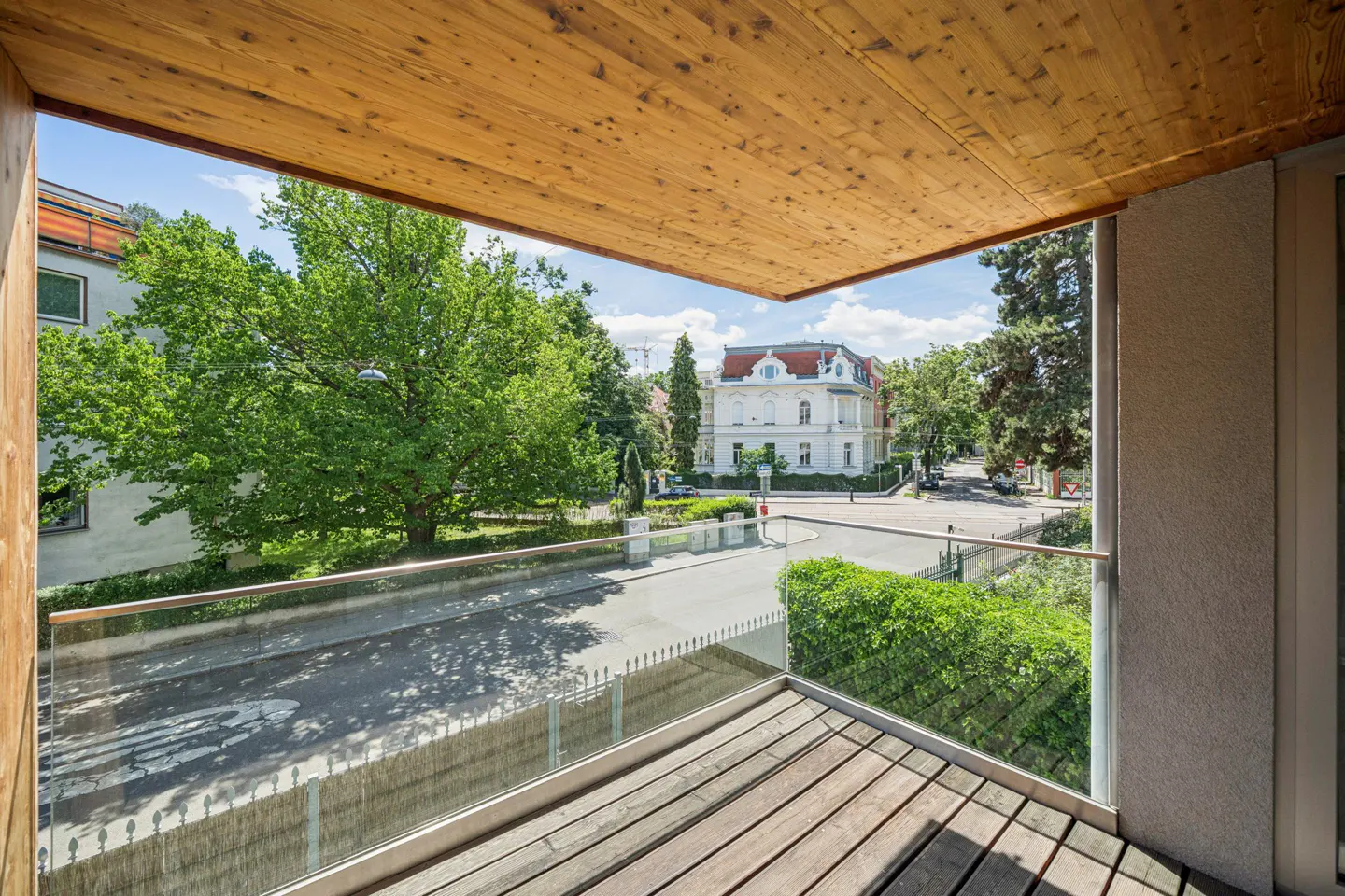 View from a modern balcony with wood ceiling and flooring, overlooking a quiet street, green trees, and a historic building.