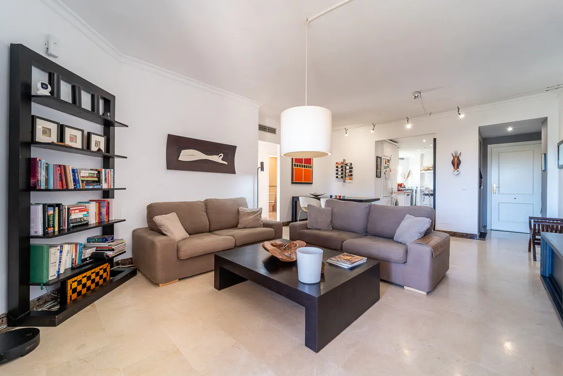 Living room with beige sofas, a dark wood coffee table, and a black bookshelf filled with books and decorative items. White walls and marble floors.