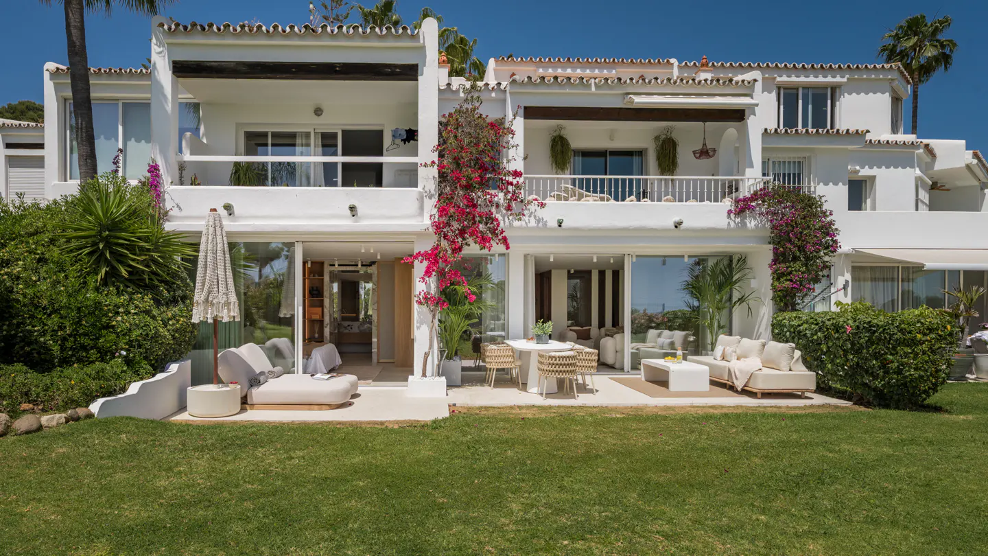 White Mediterranean villa with a lush green lawn, patio furniture, and vibrant pink bougainvillea against a blue sky.