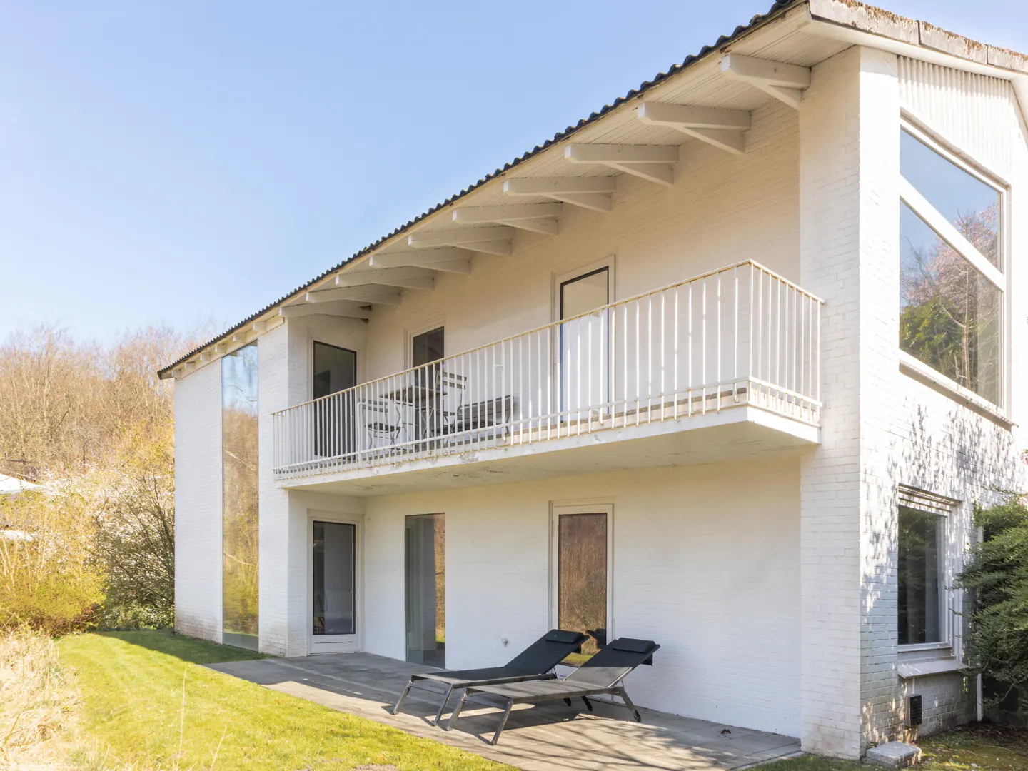 Two-story white house with a balcony and two black lounge chairs on a patio. Green grass and trees are visible.