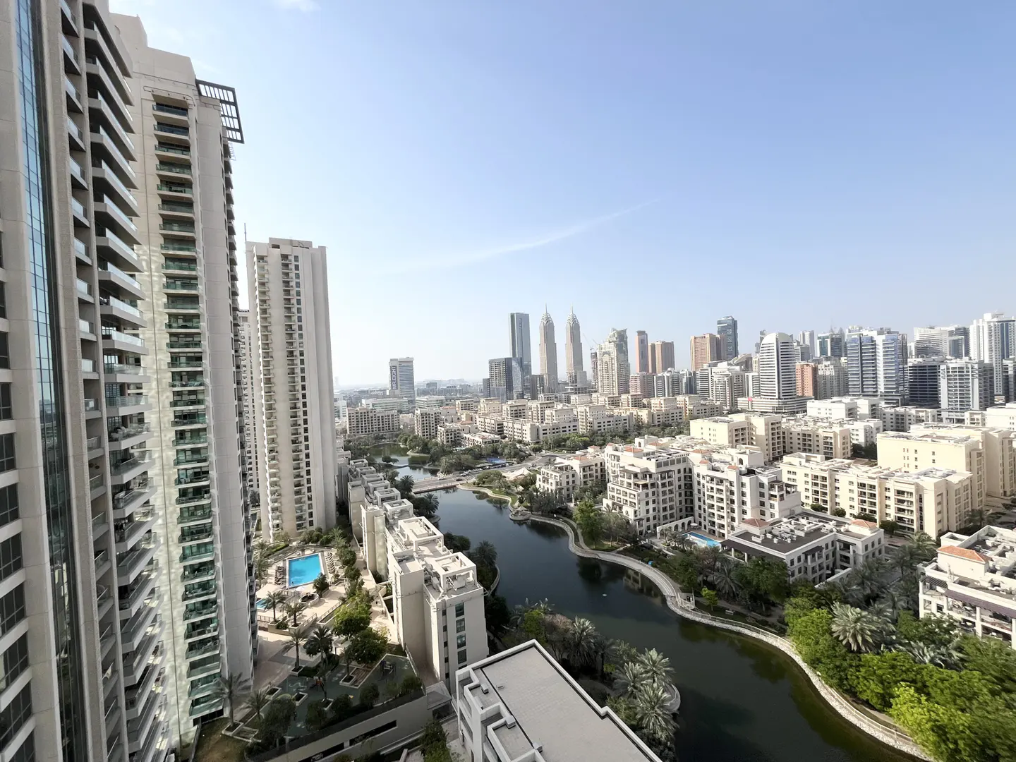 View of Dubai skyline with lake, buildings, and blue sky. Tall white apartment buildings are in the foreground.