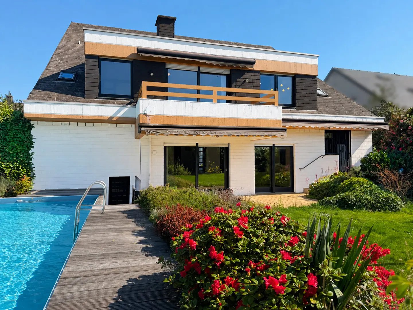 A two-story house with a pool, wooden deck, and red flowers in the front yard. The house has a balcony and black framed windows.