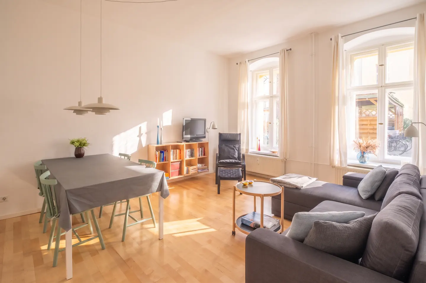 Bright living room with hardwood floors, gray sofa, dining table with green chairs, and large windows with white curtains.