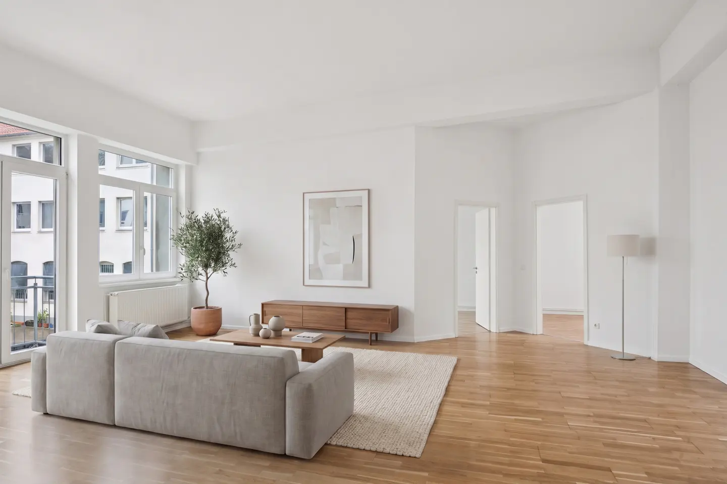 Bright, modern living room with hardwood floors, white walls, and a large gray sofa. A potted olive tree sits near the balcony doors.