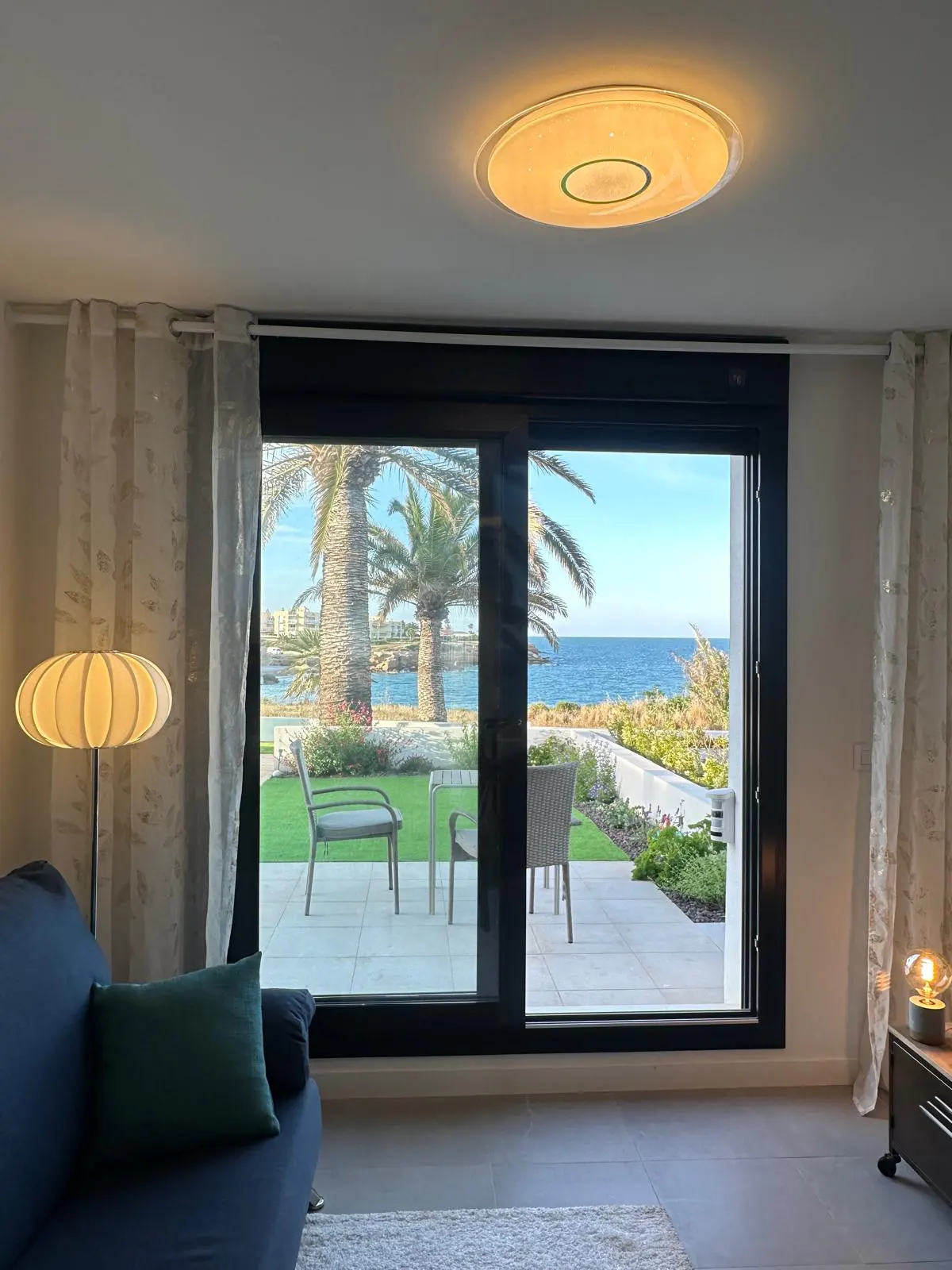 Living room view through a black-framed sliding glass door to a patio with chairs, palm trees, and the ocean.