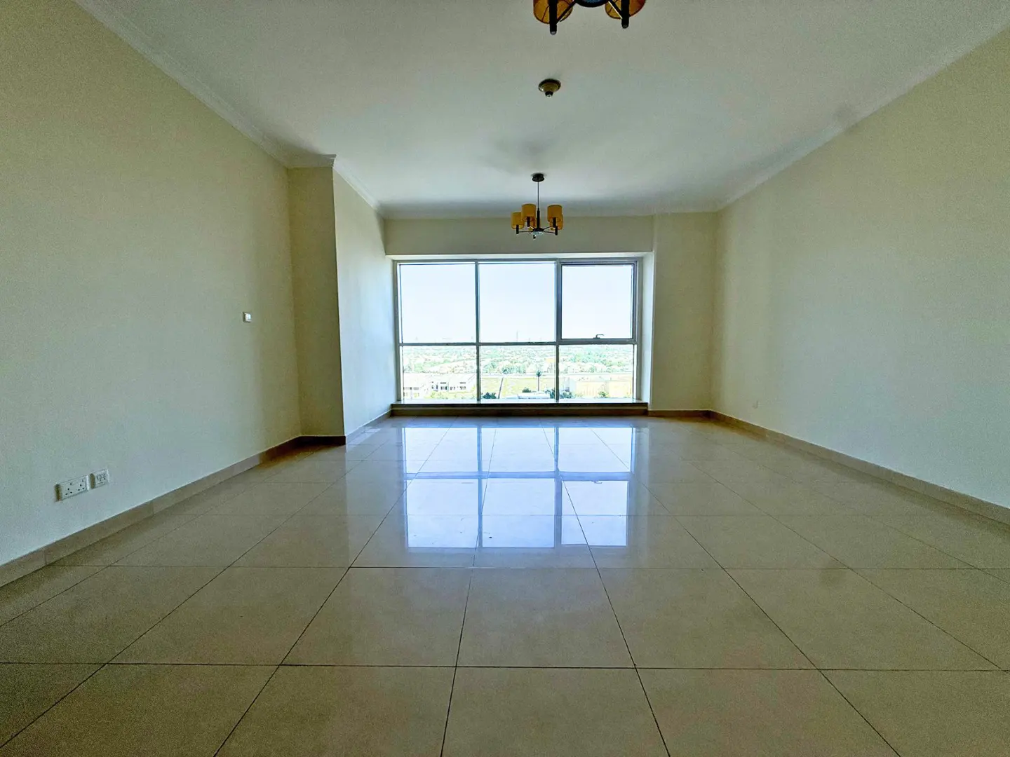 Empty room with beige walls, tile floor, and a large window with a view. Two chandeliers hang from the ceiling.