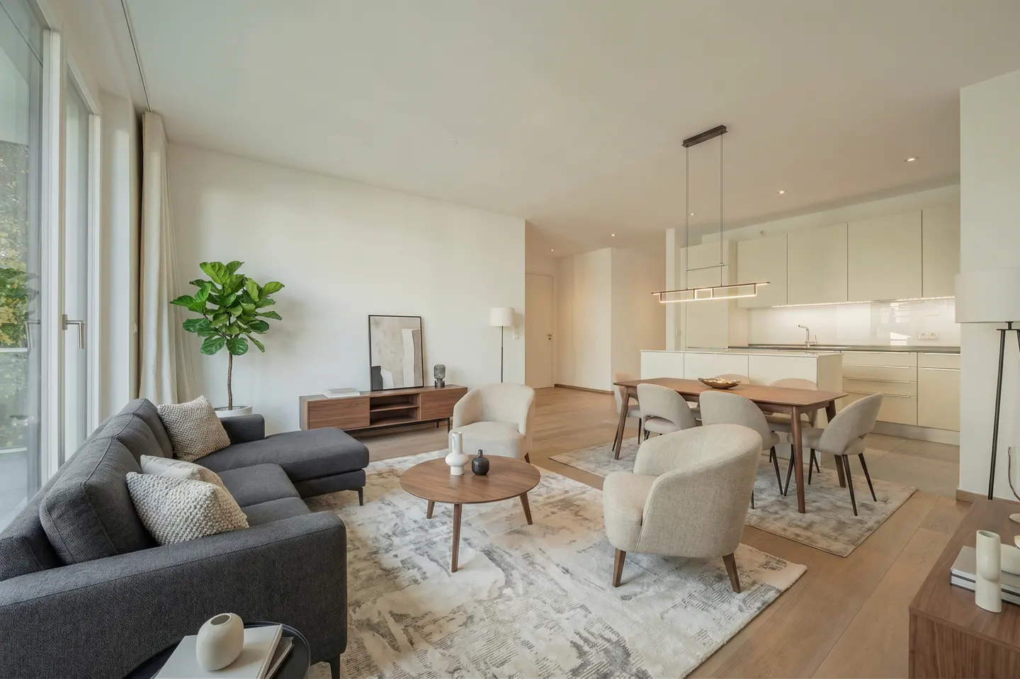 Open-concept living space with a gray sectional sofa, round wood table, and dining area with a modern light fixture.