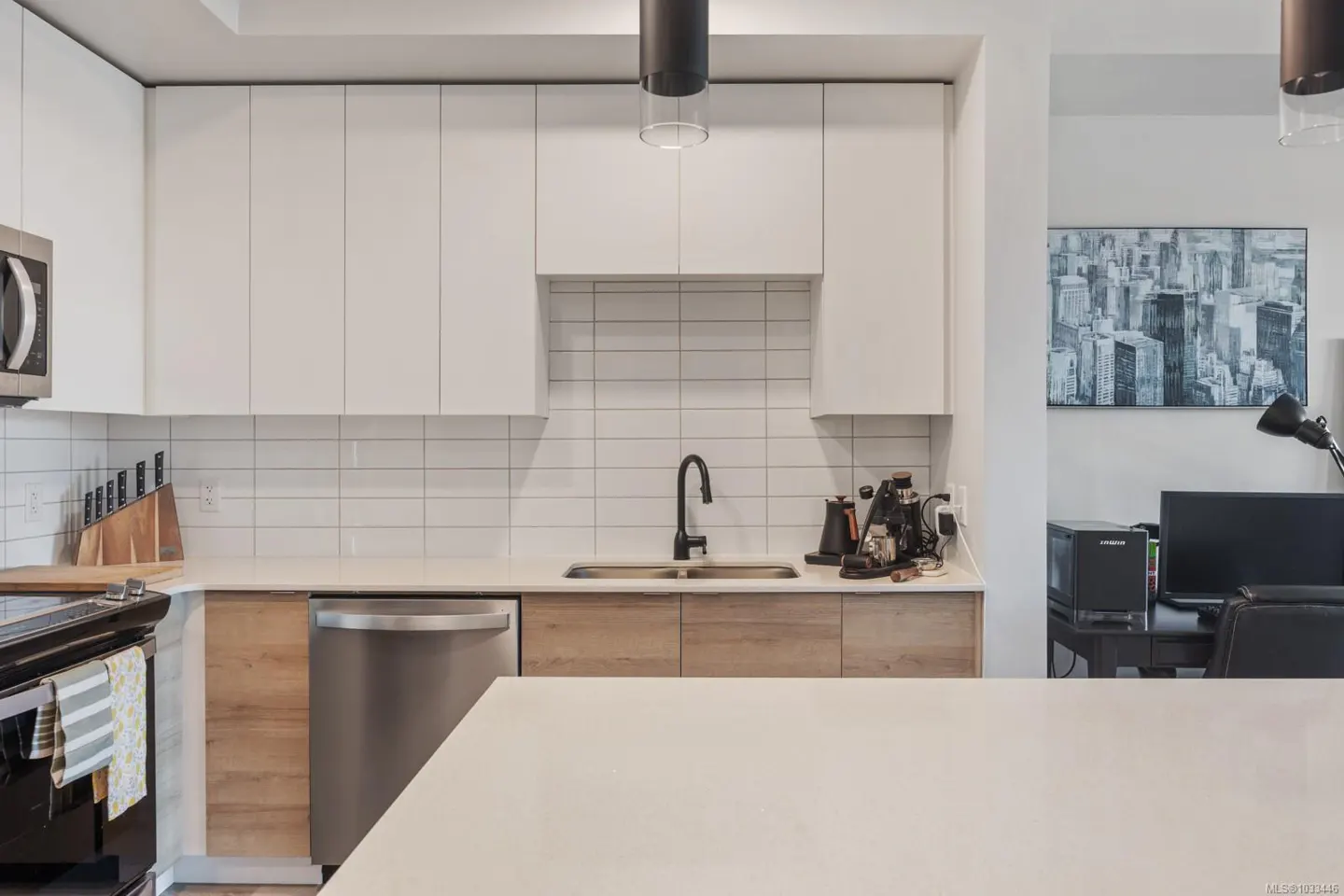 A modern kitchen with white cabinets, wood-grain lower cabinets, stainless steel appliances, and a white countertop island. A black faucet and coffee maker are visible.