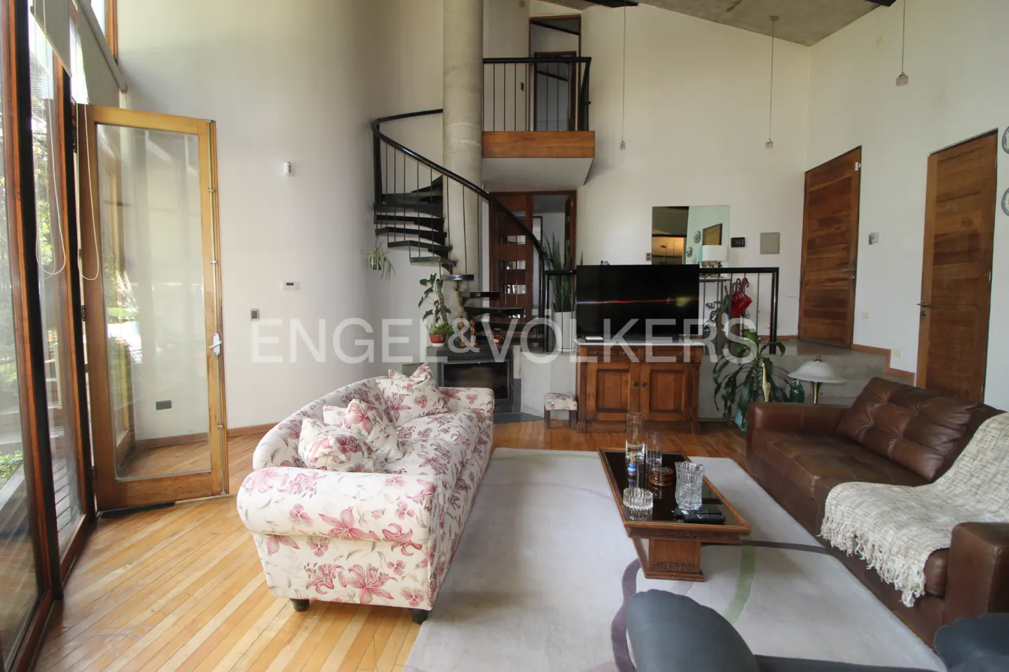 A living room with a floral sofa, brown leather sofa, spiral staircase, and wooden floors.