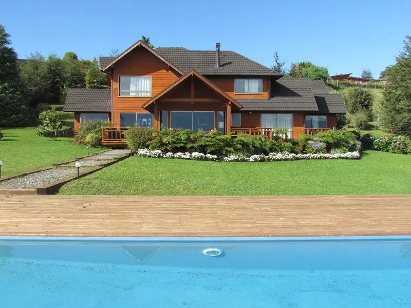 A two-story wood house with a brown roof overlooks a blue swimming pool and green lawn.