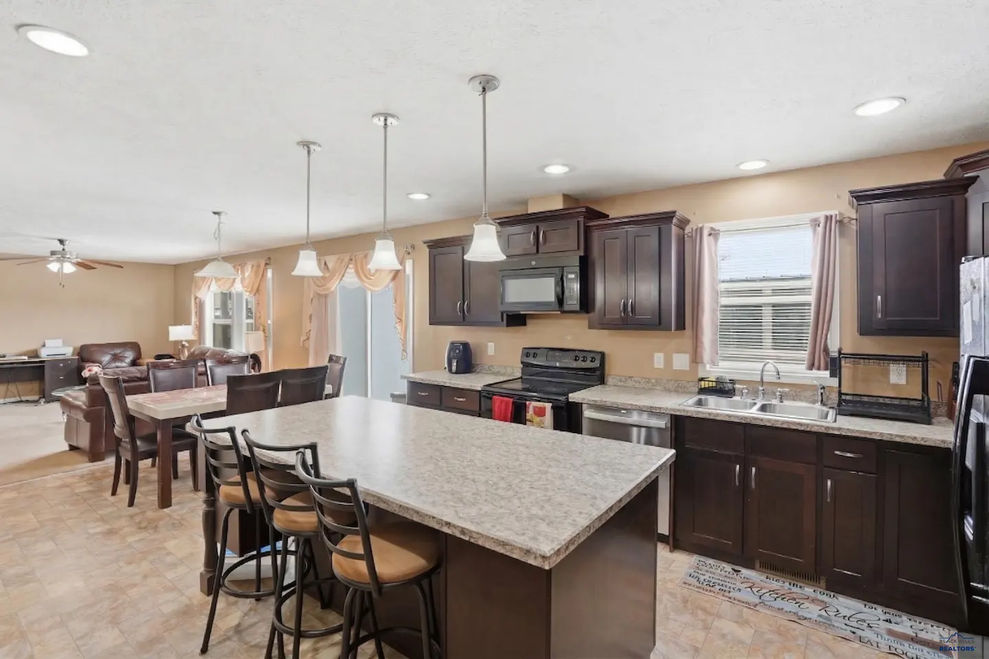 Open concept kitchen with dark cabinets, granite countertops, and island with bar stools. Living room visible in background.