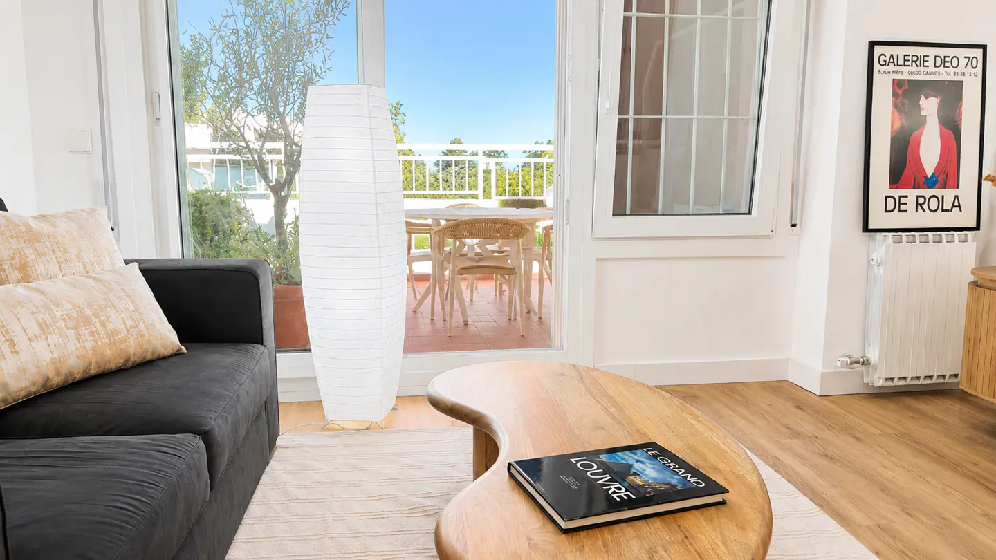 Living room with a gray sofa, wooden coffee table with a book, and a white paper lantern. Balcony with table and chairs visible through glass doors.