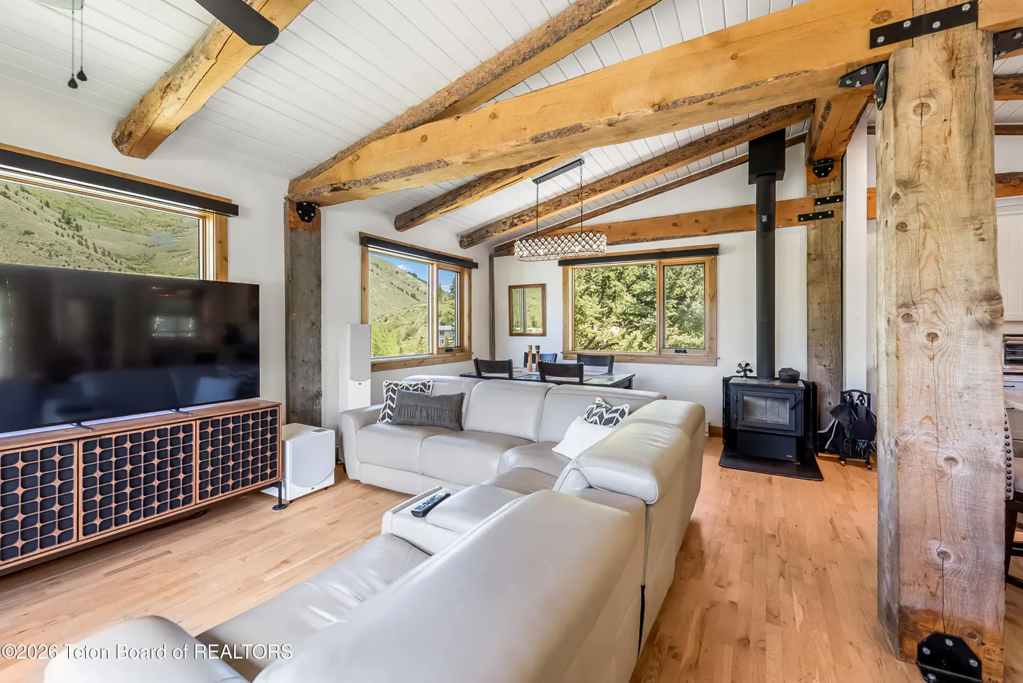 Rustic living room with vaulted ceilings, exposed wood beams, a white sectional sofa, wood stove, and mountain views.