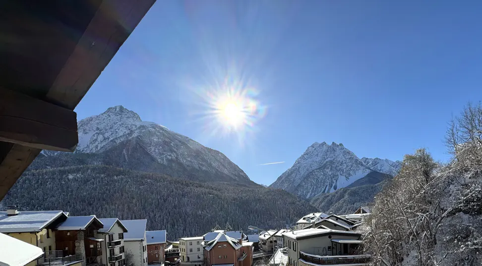 View of snow-covered mountains and houses under a bright sun and blue sky, seen from a wooden balcony.