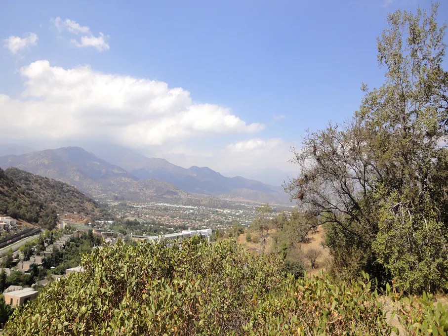 Scenic view of a valley with a town, mountains, and a blue sky with white clouds. Green trees and bushes in the foreground.