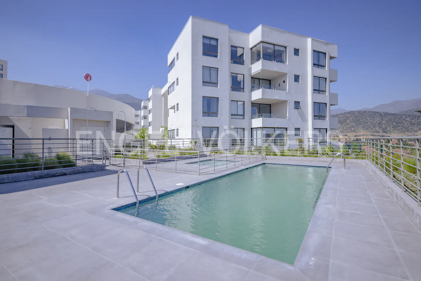 A modern white apartment building with a pool on a sunny day. Mountains are visible in the background.