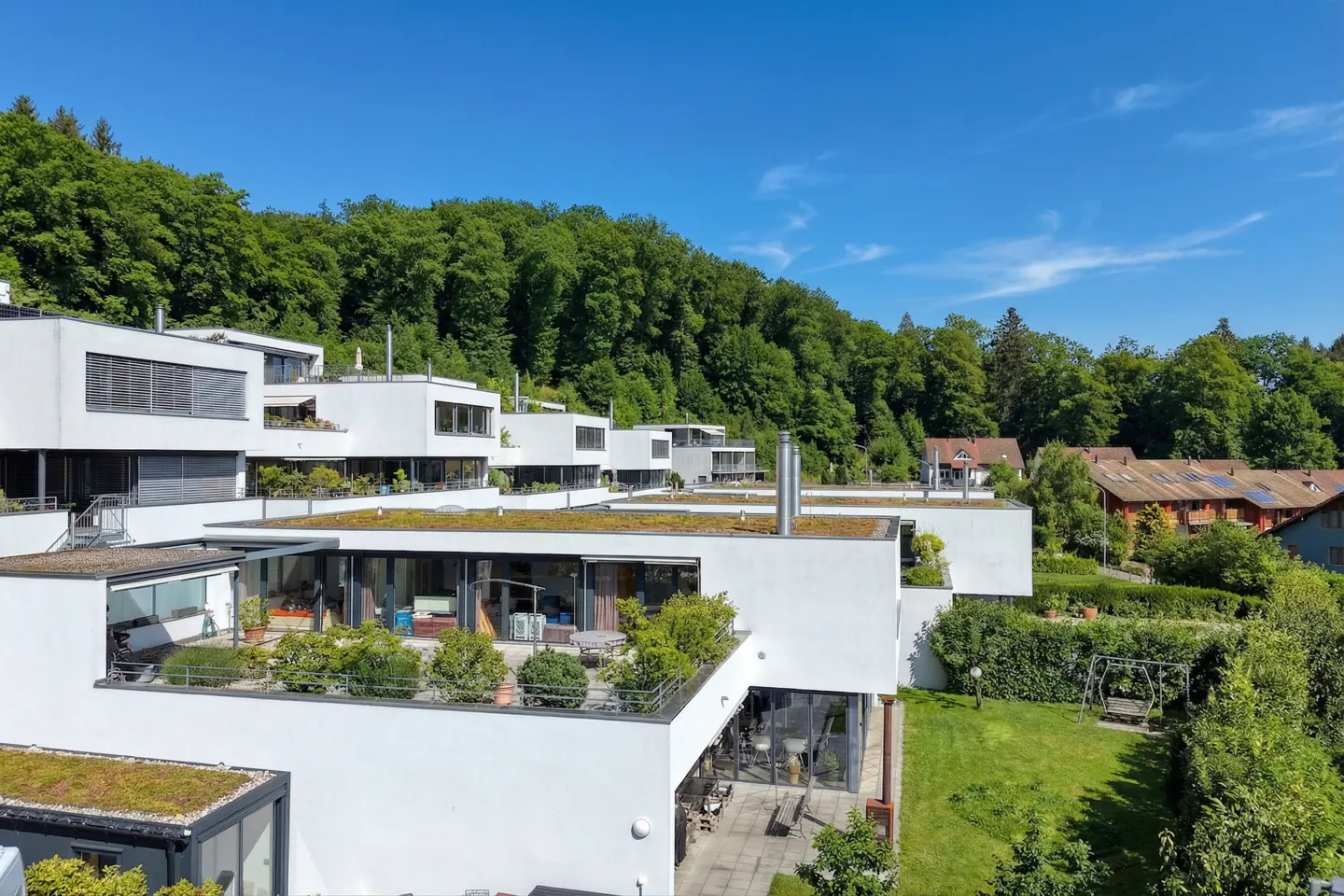 Modern white condos with green roofs cascade down a hillside under a clear blue sky. Lush green trees fill the background.