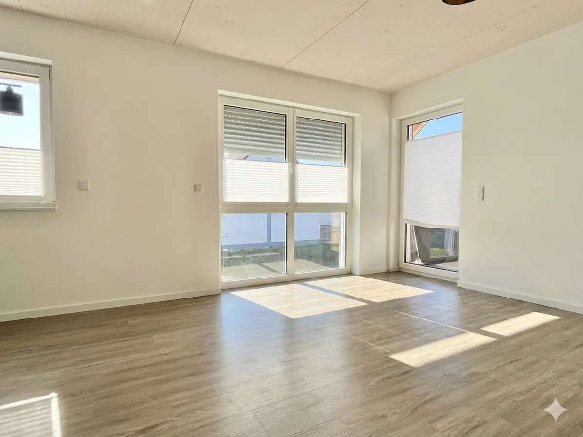 Bright, empty room with wood-look floors, white walls, and large windows with blinds. Sunlight streams across the floor.