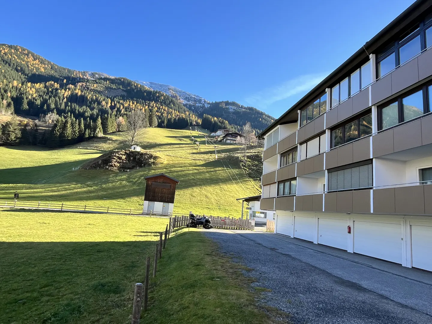 Apartment building with garages, set against a backdrop of green hills and mountains under a clear blue sky.