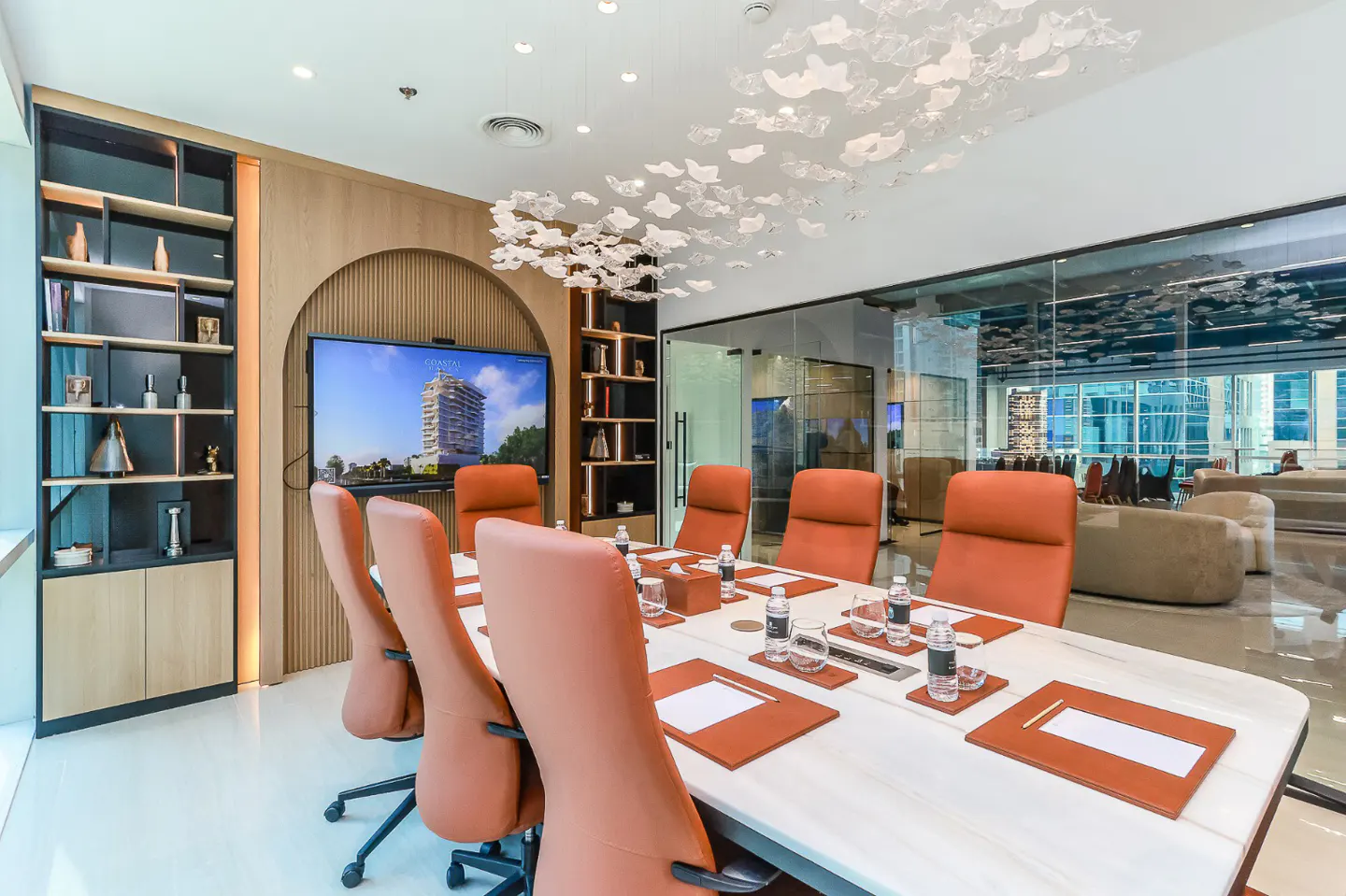 Conference room with a long white table, brown leather chairs, and a modern chandelier. A TV displays a building image.