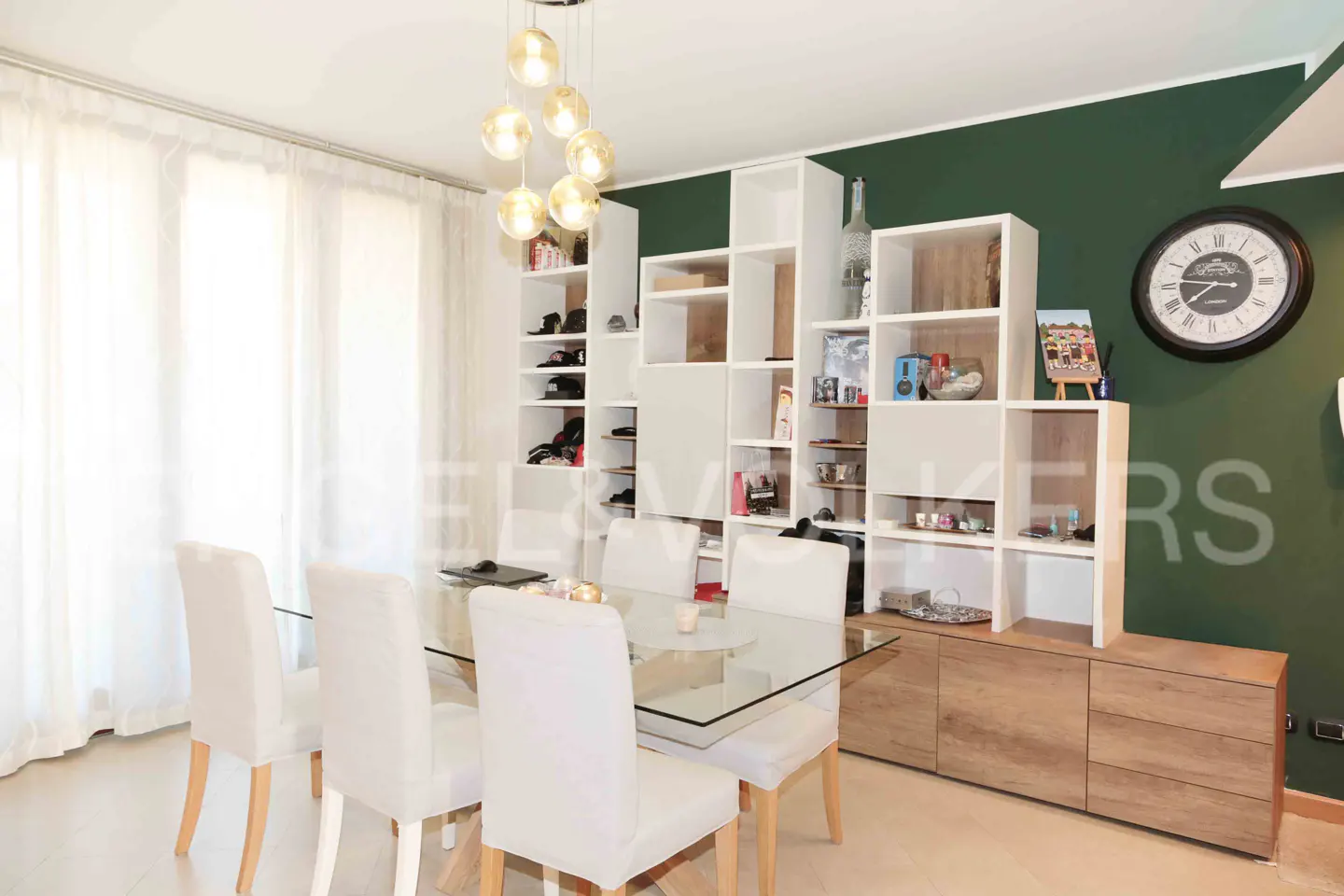 Dining room with glass table, white chairs, and a modern white bookshelf against a dark green wall. A gold chandelier hangs above.