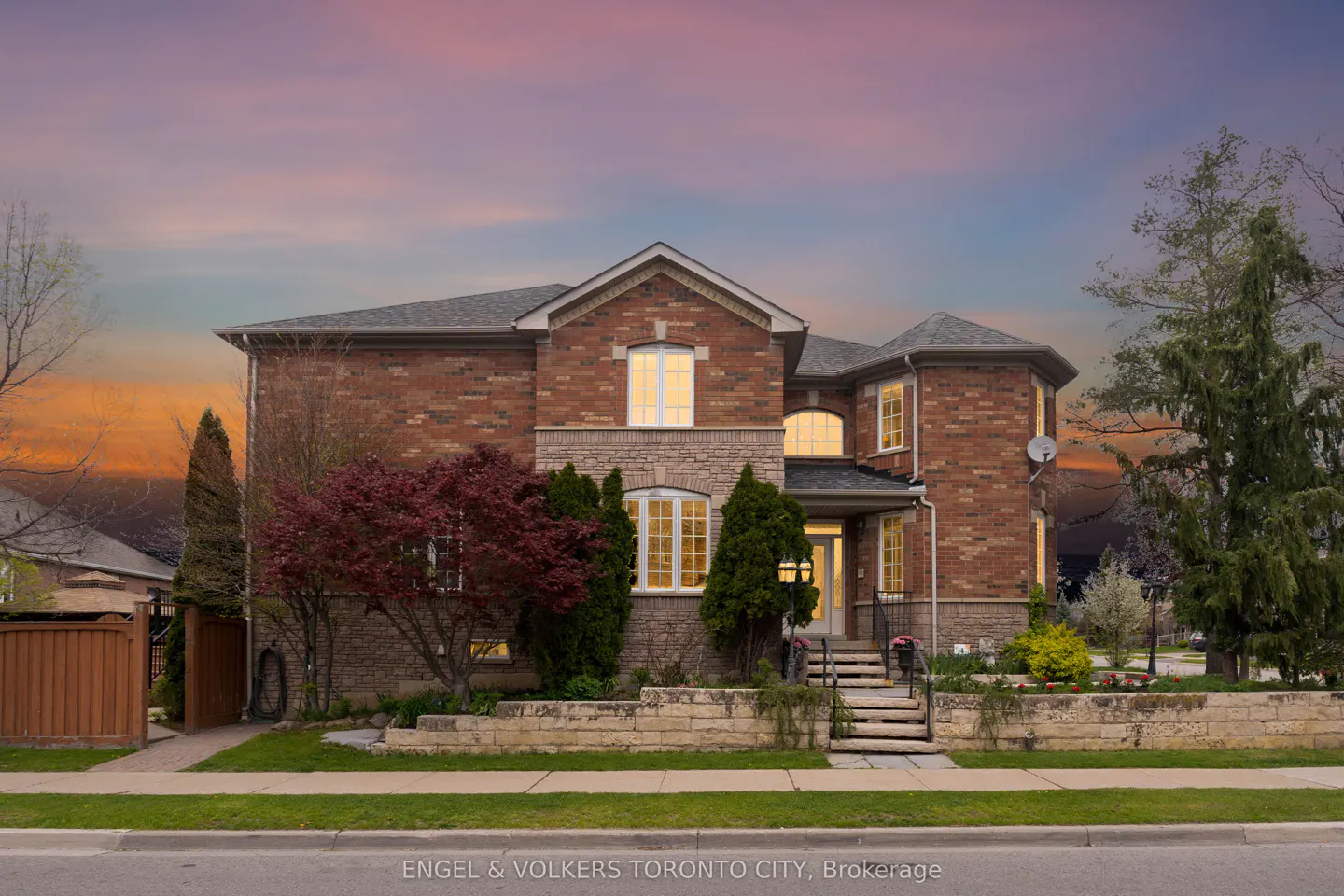 Two-story brick house with stone accents, gray roof, and illuminated windows at dusk. Landscaped front yard with trees and stone retaining wall.