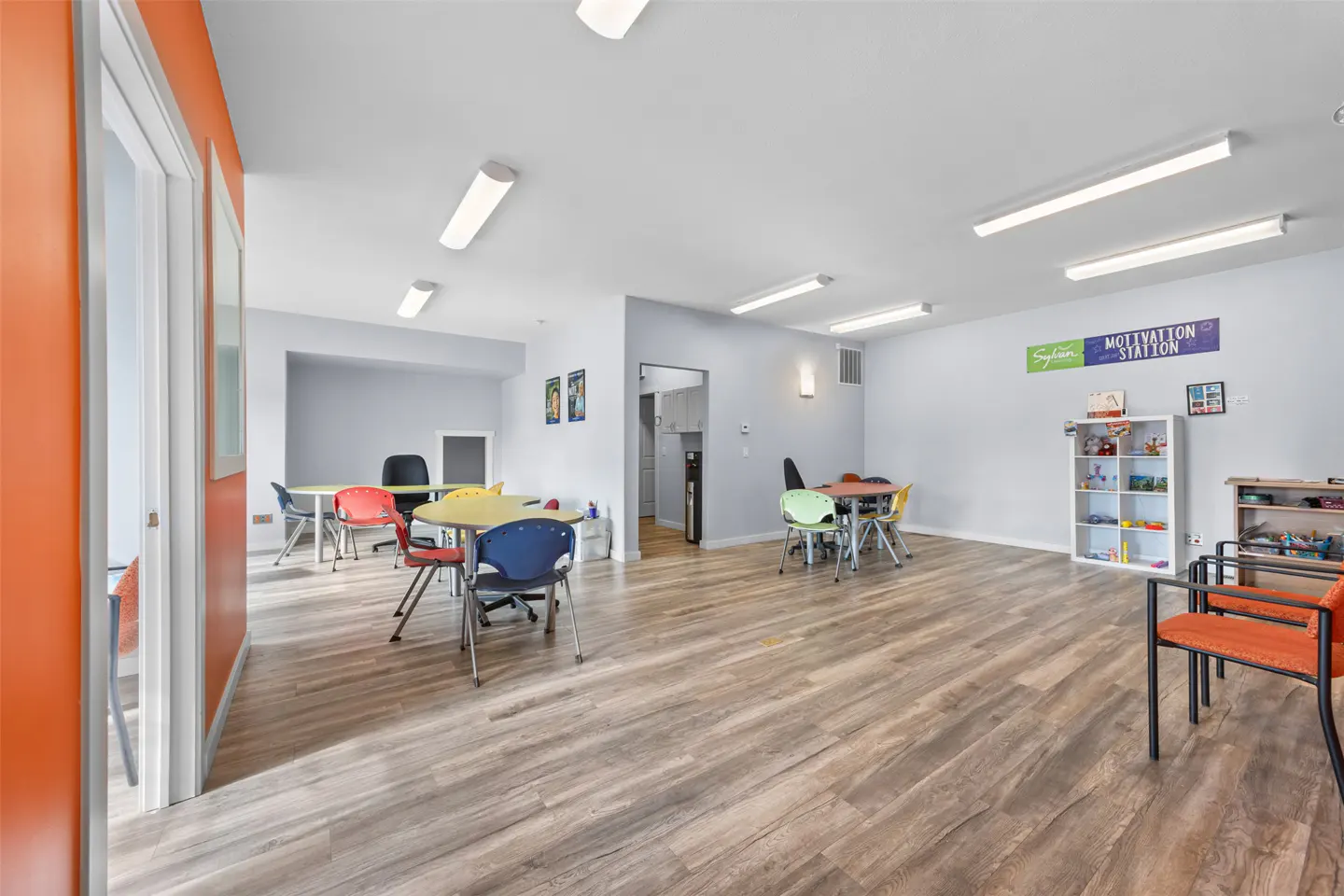 Bright room with wood floors, colorful tables and chairs, white walls, and orange accent wall. "Motivation Station" sign on the wall.