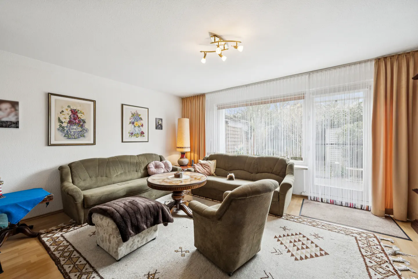 Living room with olive green sofas, armchair, and patterned rug. A round table sits in the center, with art on the white walls. Natural light streams through the window.