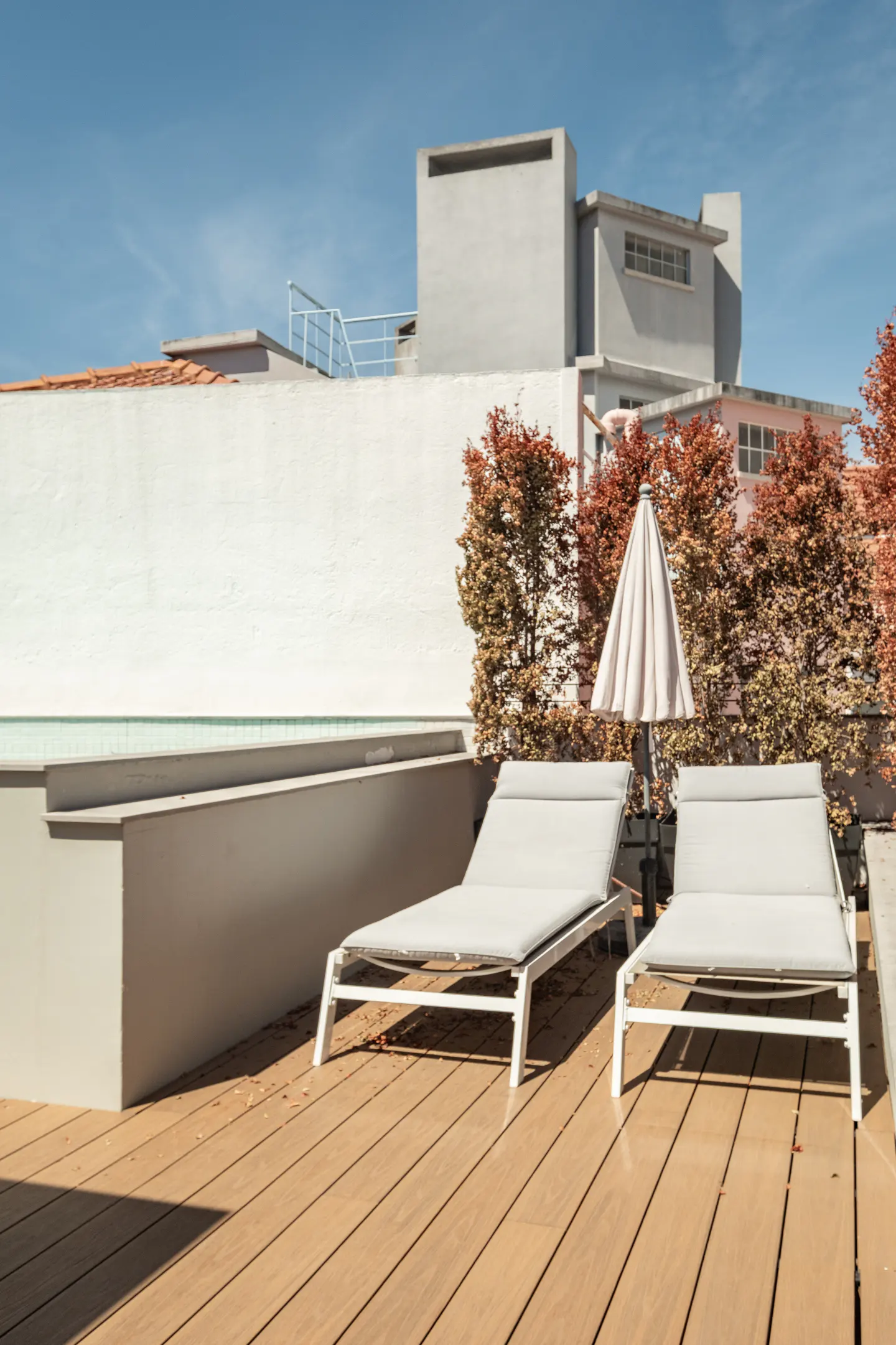 Two white lounge chairs on a wooden rooftop deck with a beige umbrella, set against a white wall and blue sky.