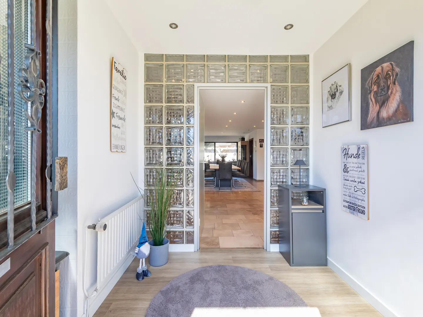 Bright entryway with white walls, wood floors, and a glass block wall leading to a dining room. Dog portraits hang on the right wall.