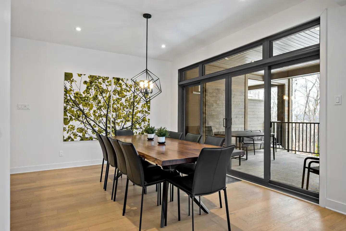 Dining room with a wood table, black chairs, and a modern light fixture. Sliding glass doors lead to an outdoor patio. A tree art piece hangs on the wall.