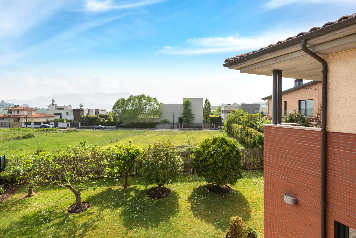 View from a balcony overlooking a green lawn with trees, houses, and a blue sky.