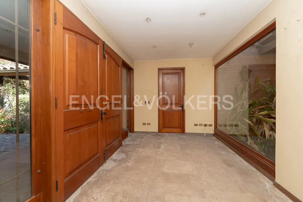 Entryway with large wooden doors, beige walls, and a large window with plants. The floor is tiled.