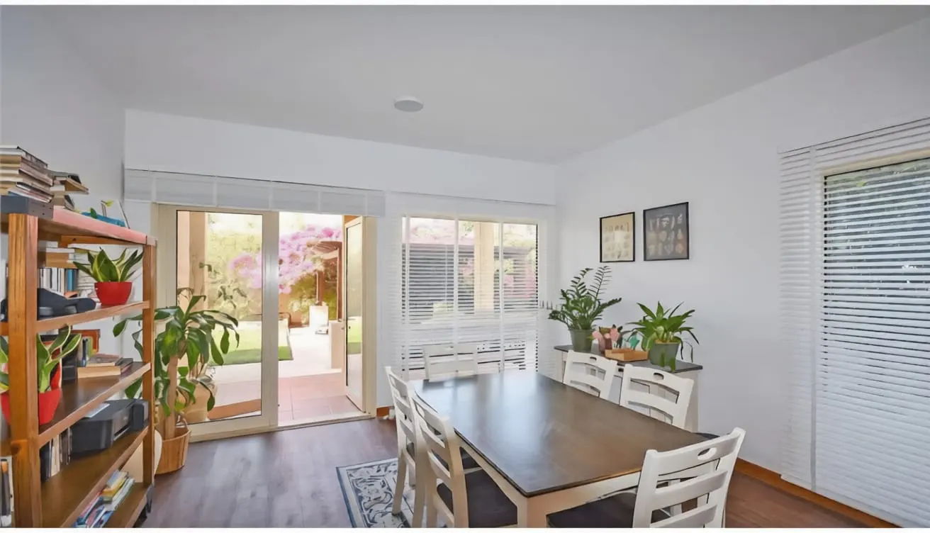 Dining room with a dark wood table, white chairs, potted plants, and a glass door opening to a sunny patio with flowers.