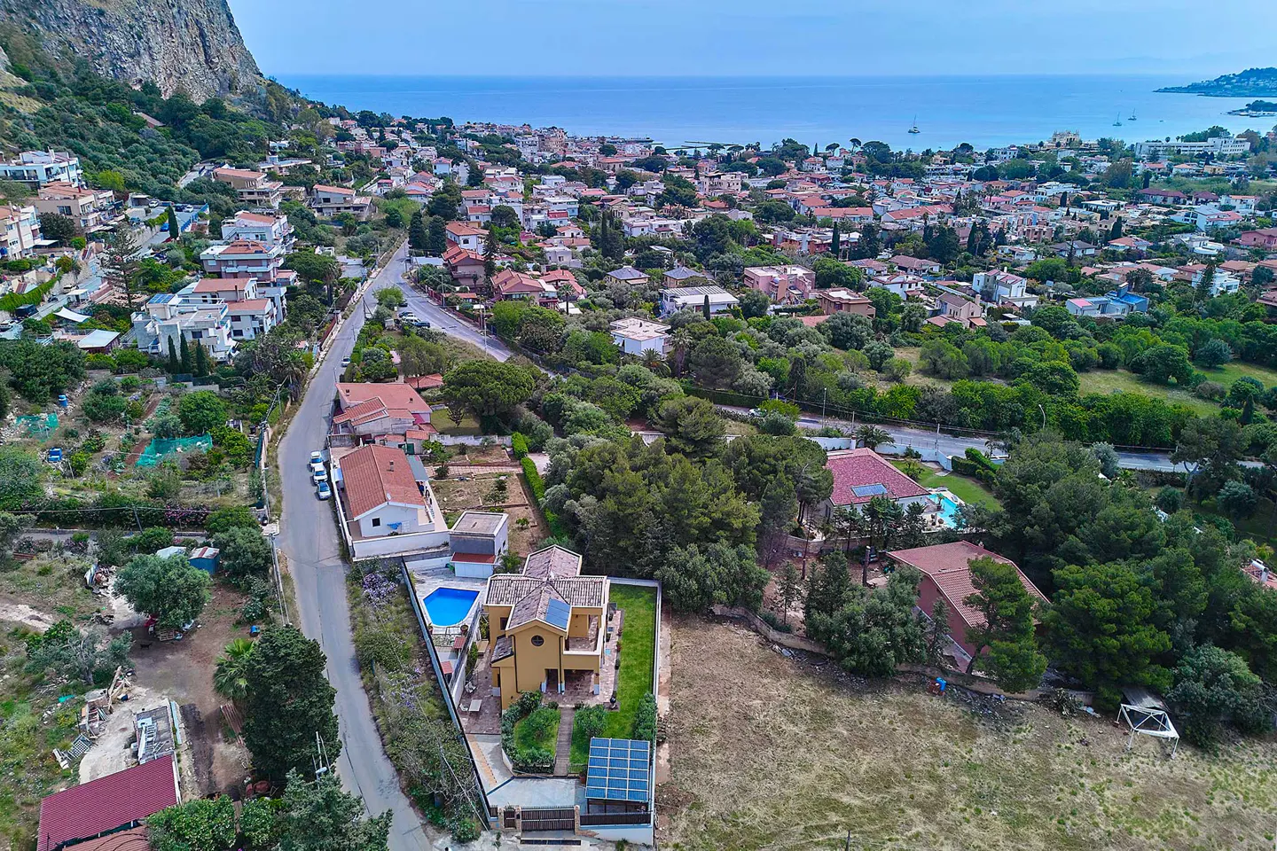 Aerial view of a yellow house with a blue pool, nestled among green trees in a coastal town with the ocean in the background.