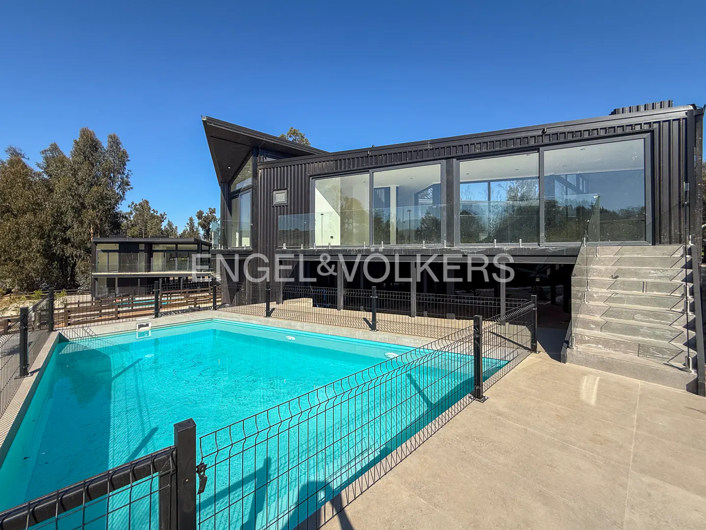 Modern black house with a pool. Large windows reflect the sky. A black fence surrounds the pool. Trees are in the background.
