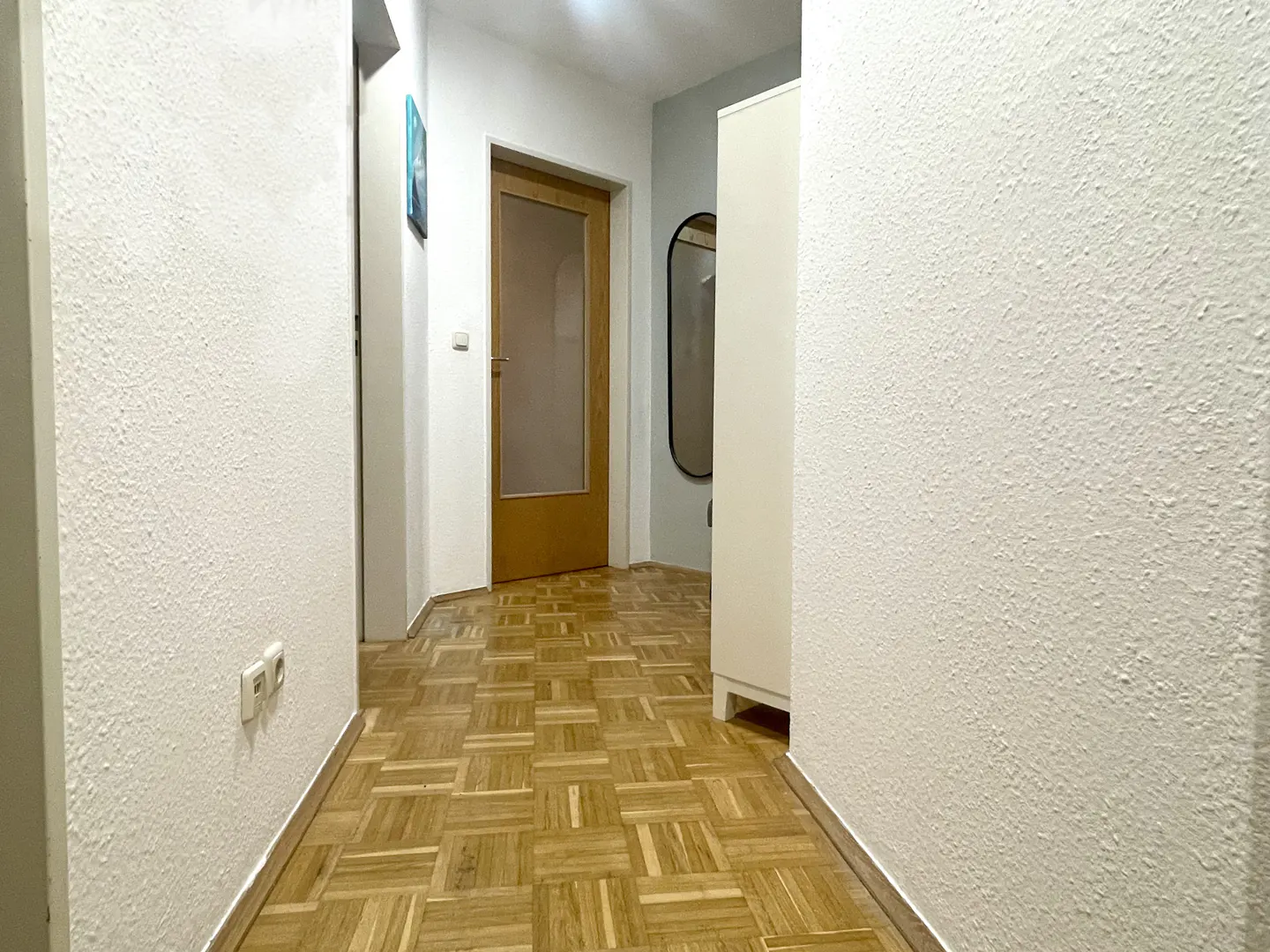Hallway with parquet flooring, white textured walls, and a wooden door with glass panel. A white wardrobe and oval mirror are visible.