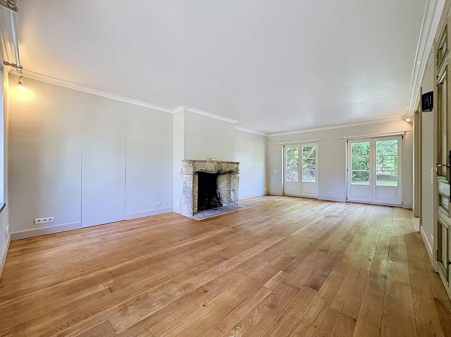 Bright, empty living room with hardwood floors, a stone fireplace, and white French doors leading to a green outdoor space.