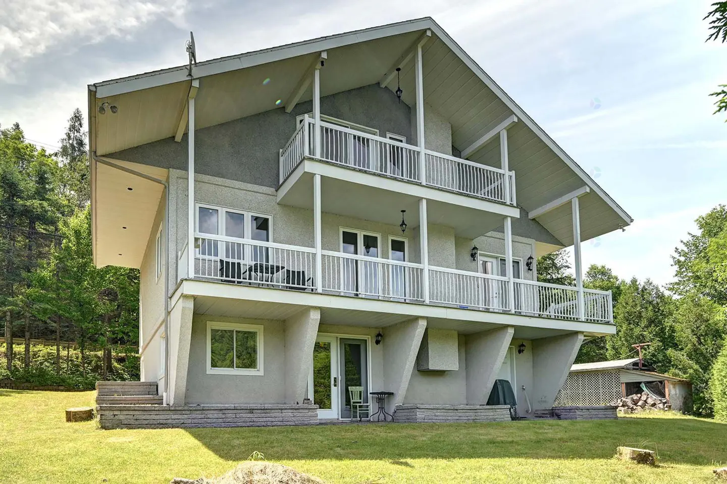 Two-story house with white balconies and gray siding, surrounded by green trees and grass.