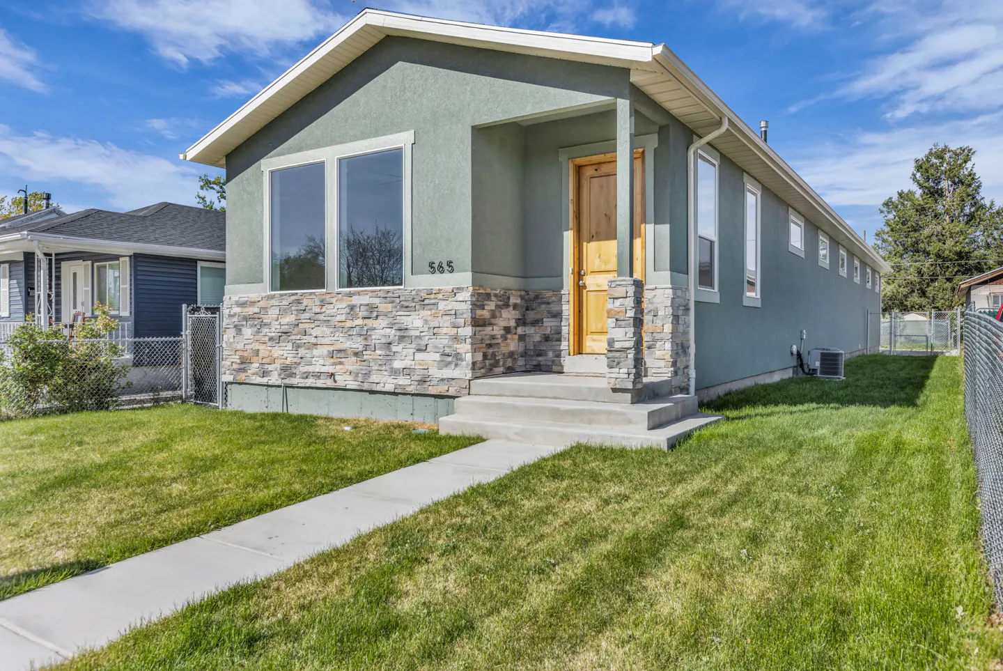 Exterior of a one-story, sage green house with stone accents, a wooden front door, and a concrete walkway leading to the entrance.