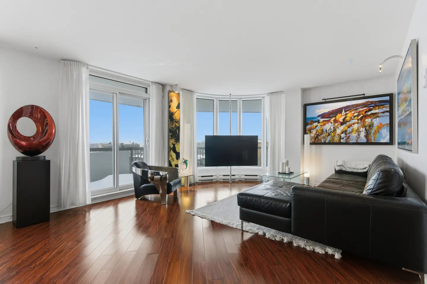 Bright living room with hardwood floors, black leather sofa, modern art, and large windows with city views.