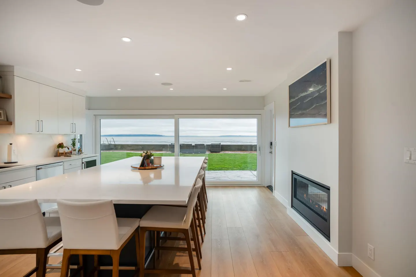 Bright, modern kitchen with a large island and white cabinets. A sliding glass door leads to a green lawn and ocean view. Fireplace on the right wall.
