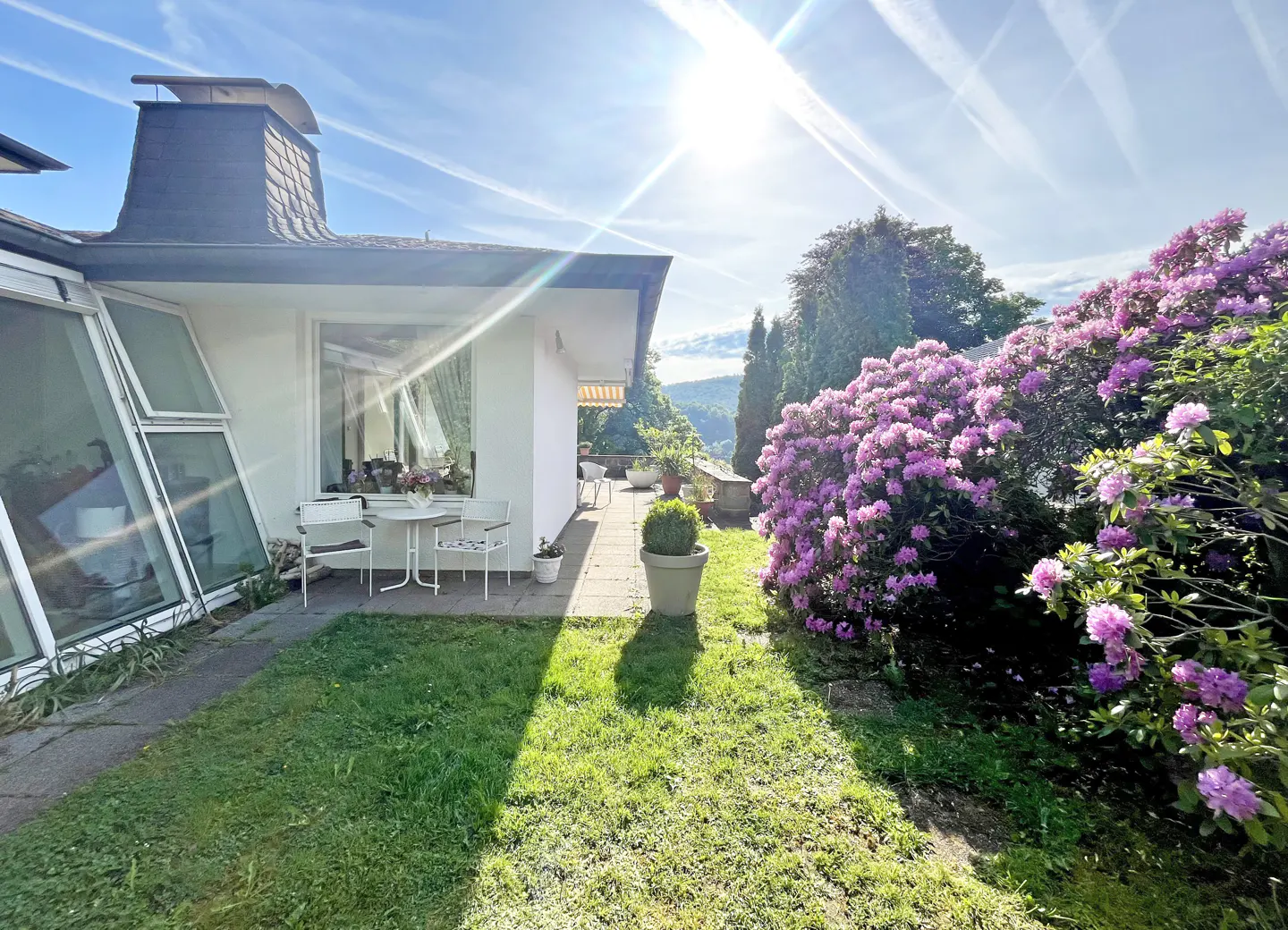 Sunny patio with white table and chairs, green lawn, and blooming purple rhododendrons.